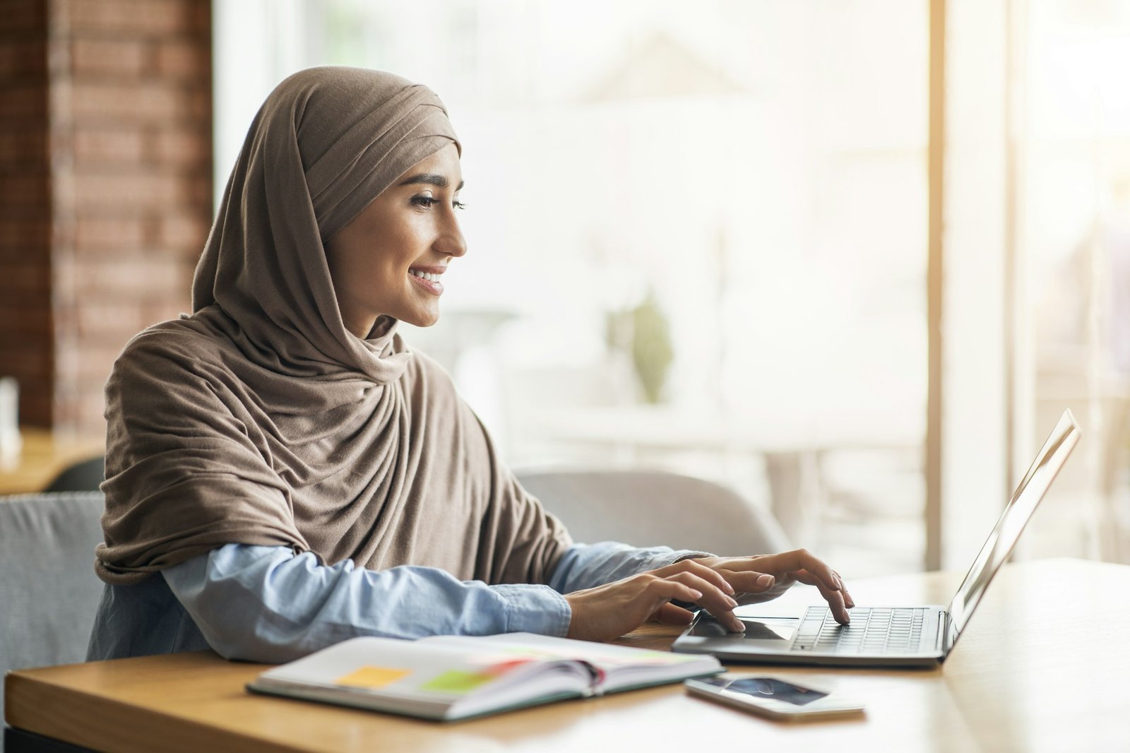 Cheerful girl in hijab sitting at cafe, looking for job